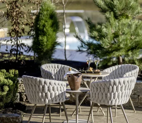 Terrasse einer NaturLodge des Sonnenalp Resorts mit vier gepolsterten Stühlen, gedecktem Tisch, Champagner und Snacks, umgeben von Pflanzen und Pool im Hintergrund