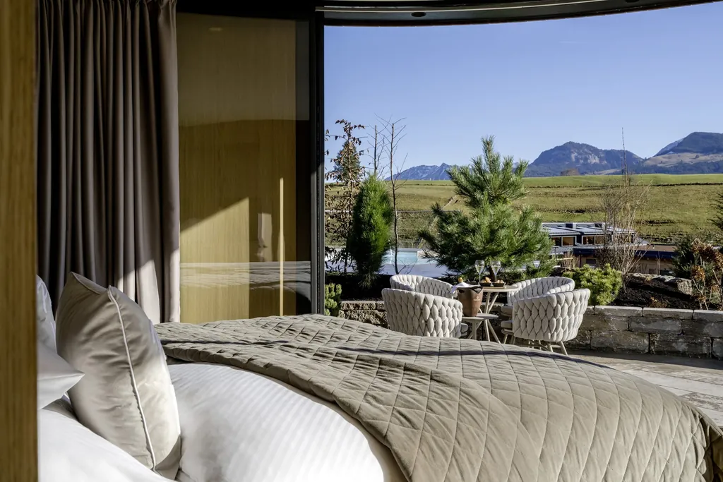 Schlafzimmer einer NaturLodge des Sonnenalp Resorts mit einem Doppelbett und Blick auf eine Terrasse mit Sitzmöbeln, Natur und Berge im Hintergrund