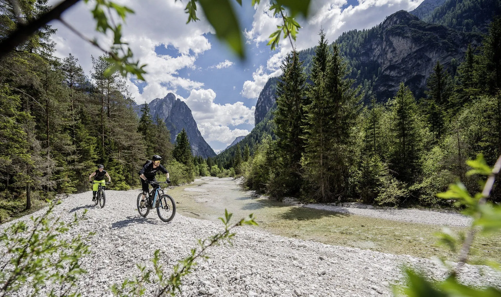 Zwei Mountainbiker fahren auf einem steinigen Weg entlang eines klaren Bachs, umgeben von gr&uuml;nen Nadelw&auml;ldern und imposanten Berggipfeln in der N&auml;he des Hotel Winkler
