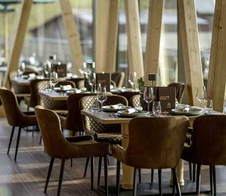 Gedeckte Tische mit braunen Polsterstühlen im Hofgut-Restaurant des Sonnenalp Resorts, mit großen Holzsäulen und Blick nach draußen