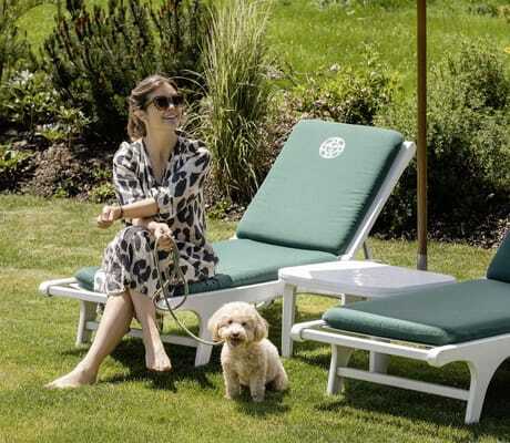 Frau mit Hund an der Leine sitzt auf einer Liege mit grünem Polster auf einer gepflegten Wiese im Sonnenalp Resort