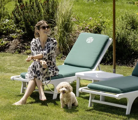 Frau mit Hund an der Leine sitzt auf einer Liege mit grünem Polster auf einer gepflegten Wiese im Sonnenalp Resort