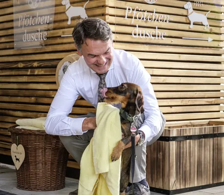 Mann trocknet einen kleinen Hund vor einer Holzfassade mit der Beschriftung „Pfötchendusche“ im Sonnenalp Resort