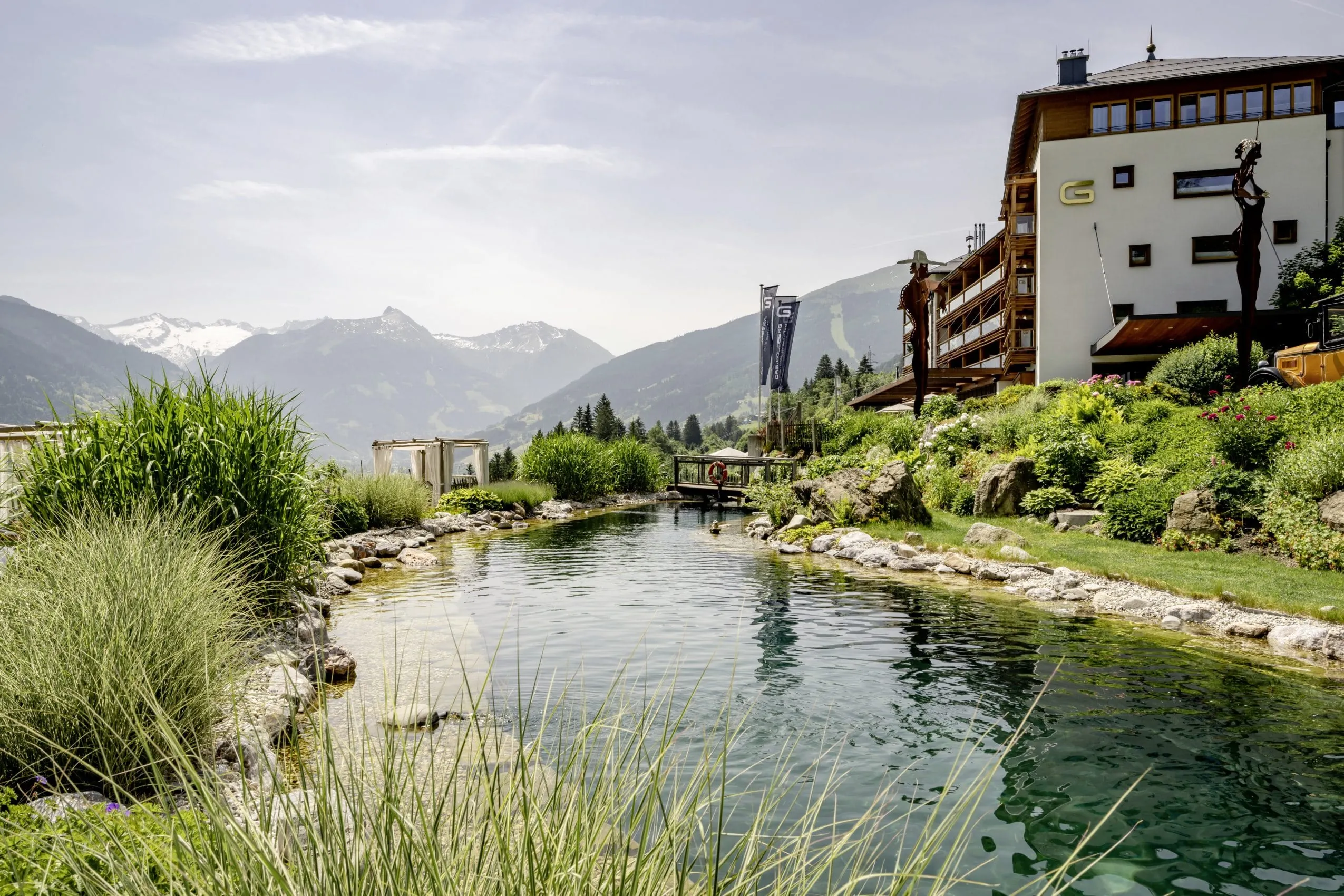 Idyllischer Naturbadesee mit Himmelbetten neben der Hotelfassade des DAS.GOLDBERG mit Bergpanorama im Hintergrund