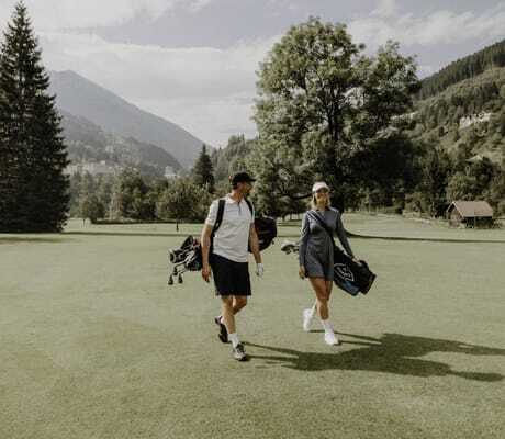 Zwei Personen mit Golfausrüstung auf einer grünen Golfwiese, umgeben von Bäumen und Bergen in der Nähe des DAS.GOLDBERG