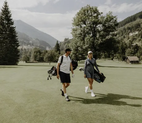 Zwei Personen mit Golfausrüstung auf einer grünen Golfwiese, umgeben von Bäumen und Bergen in der Nähe des DAS.GOLDBERG