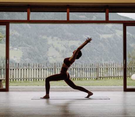 Frau beim Yoga in einer Pose vor großen Fenstern mit Bergblick und Holzboden im DAS.GOLDBERG