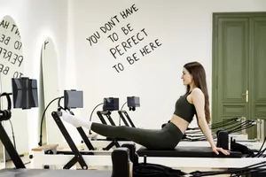 Frau in schwarzer Sportkleidung macht eine Übung auf einem Reformer-Pilates-Gerät in einem hellen Raum mit Spiegeln und Motivationsschriftzug an der Wand