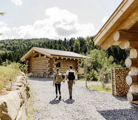 Paar mit Rucksäcken läuft Händchen haltend an den im Blockhausstil gebauten Jakob Chalets vorbei