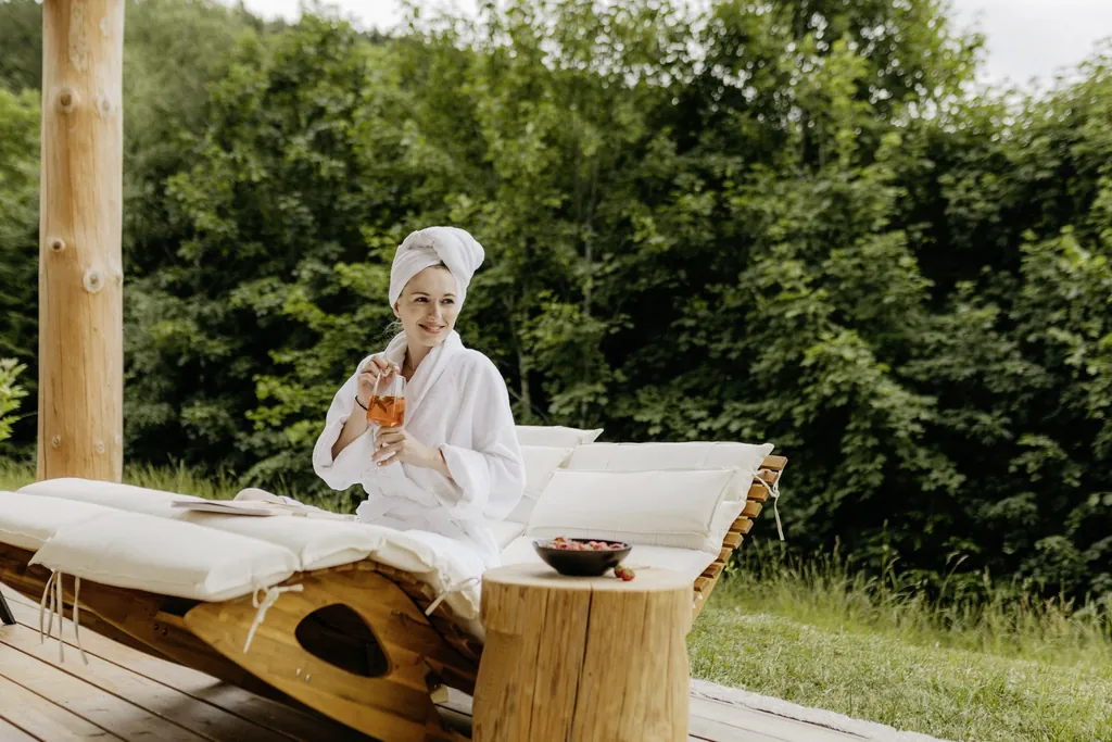 Frau in Bademantel, mit Haarturban und einem Aperitif auf einer geschwungenen Holzliege auf der Terrasse der Jakob Chalets
