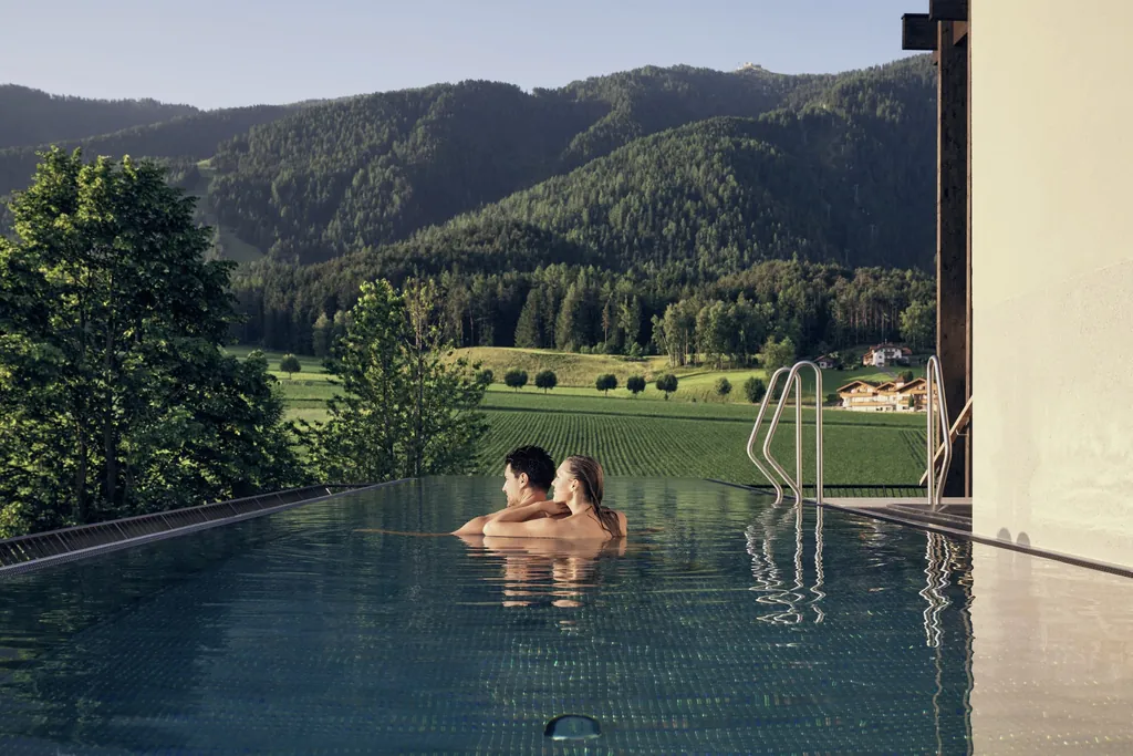 Ein Paar im Rooftop-Pool des Falkensteiner Hotel Kronplatz mit Blick auf grüne Wälder und Wiesen.