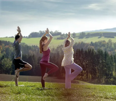 Drei Personen praktizieren Yoga im Freien mit grünen Hügeln im Hintergrund, unweit vom Landrefugium Obermüller in Untergriesbach.