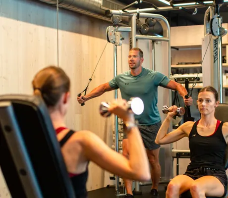 Moderner Fitnessraum mit einer Frau, die Hanteln hebt und einem Mann, der an einer Kabelzugmaschine trainiert, umgeben von Holzverkleidung und weiteren Sportgeräten im Good Life Resort Riederalm