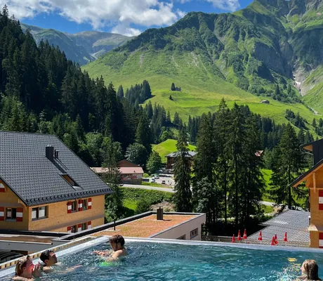 Mehrere Personen im Outdoorpool des Berghaus Schröcken mit Blick auf grüne Berghänge, umliegende Gebäude und Wald