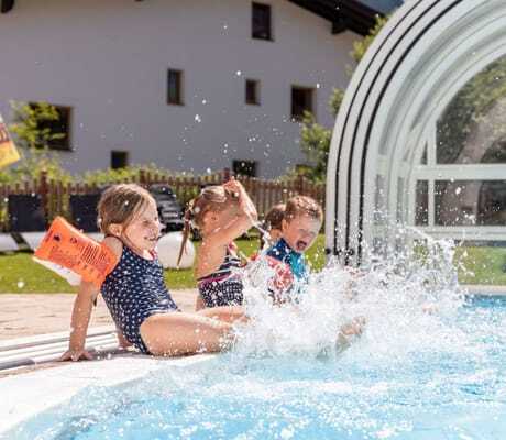 Kinder in Badeanzügen planschen fröhlich am Rand des Outdoorpools im Familotel Kaiserhof vor einer überdachten Glaskonstruktion und einem Gartenbereich.