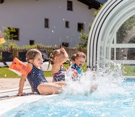 Kinder in Badeanzügen planschen fröhlich am Rand des Outdoorpools im Familotel Kaiserhof vor einer überdachten Glaskonstruktion und einem Gartenbereich.