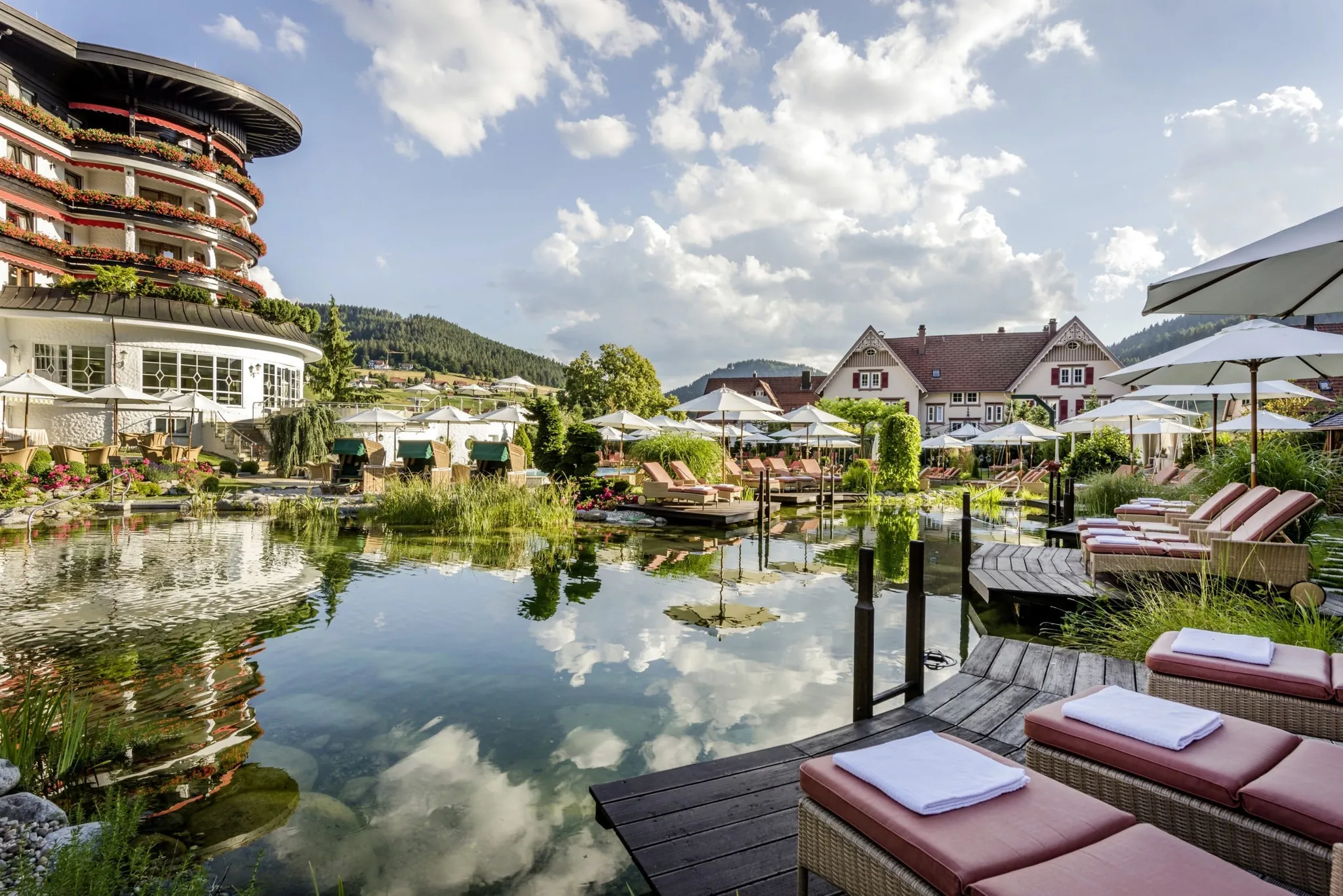 Außenansicht des Hotel Bareiss mit Naturteich im Vordergrund und einigen Sonnenliegen sowie weißen Sonnenschirmen