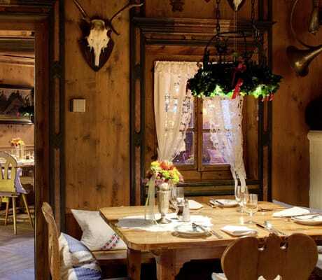 Rustikale Dorfstube im Hotel Bareiss mit Holzwänden, dekorativen Hirschgeweihen, einem gedeckten Holztisch und einem Fenster mit Spitzenvorhang
