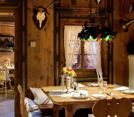 Rustikale Dorfstube im Hotel Bareiss mit Holzwänden, dekorativen Hirschgeweihen, einem gedeckten Holztisch und einem Fenster mit Spitzenvorhang