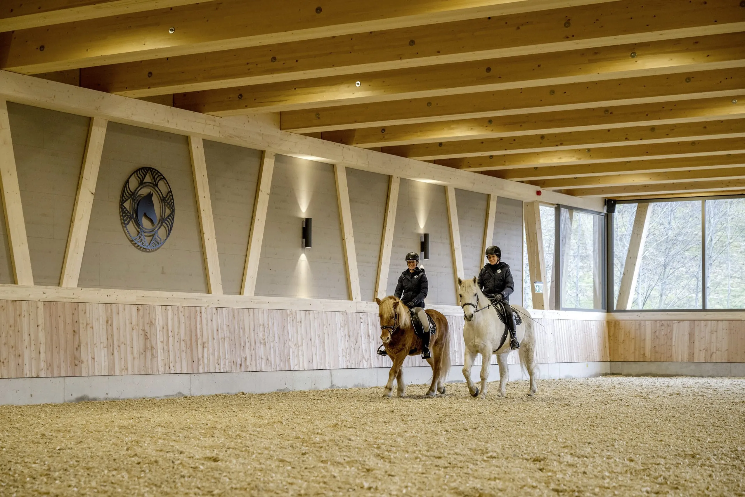 Zwei Reiter in einer modernen Holz-Reithalle des Sonnenalp Resorts auf einem braunen und einem wei&szlig;en Pferd
