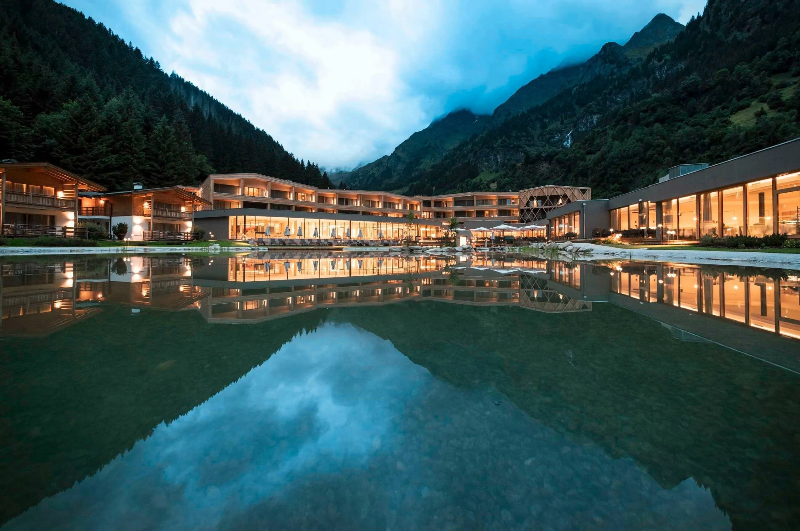Beleuchtete Außenansicht des Feuerstein Nature Family Resort mit See und grünen Berghängen und Wäldern im Hintergrund.