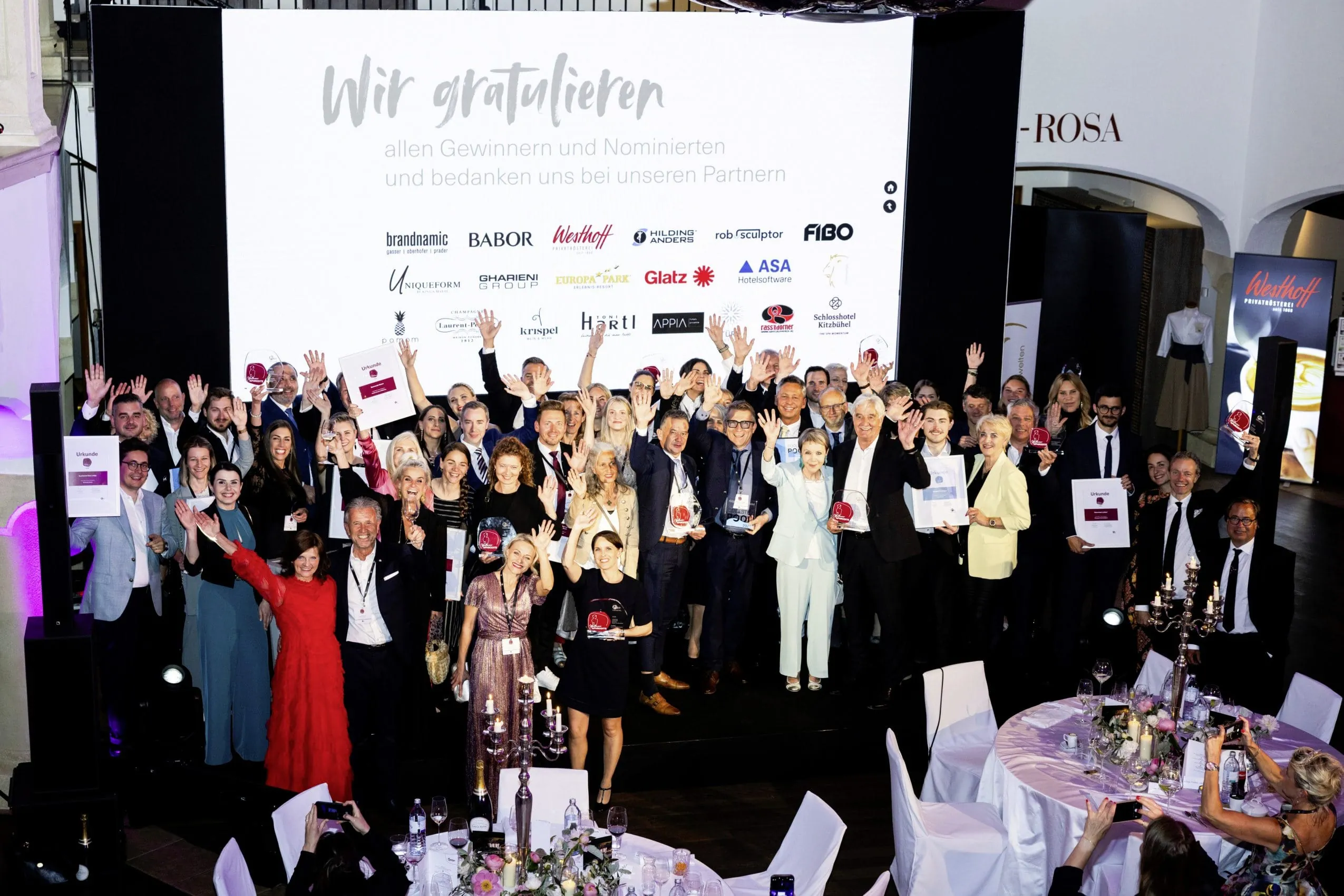 Gro&szlig;e Gruppe von Preistr&auml;gern und Teilnehmern bei der Awardverleihung Wellness Aphrodite, stehend vor einer B&uuml;hne mit Gratulationsbotschaft und Sponsoren-Logos