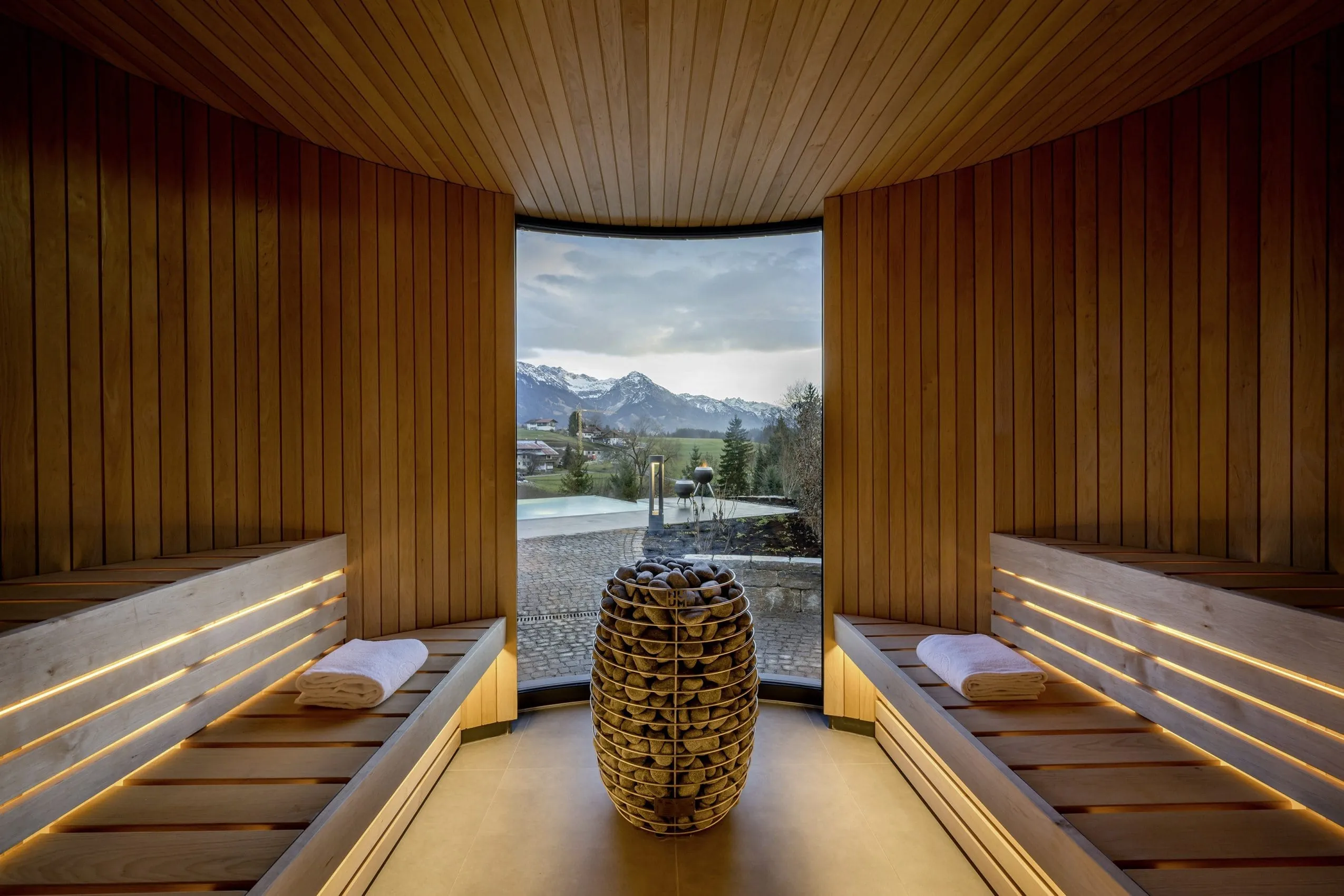 Moderne Holzsauna einer NaturLodge des Sonnenalp Resorts mit Panoramafenster und Steinofen in der Mitte, Blick auf Berge und Außenbereich mit Pool