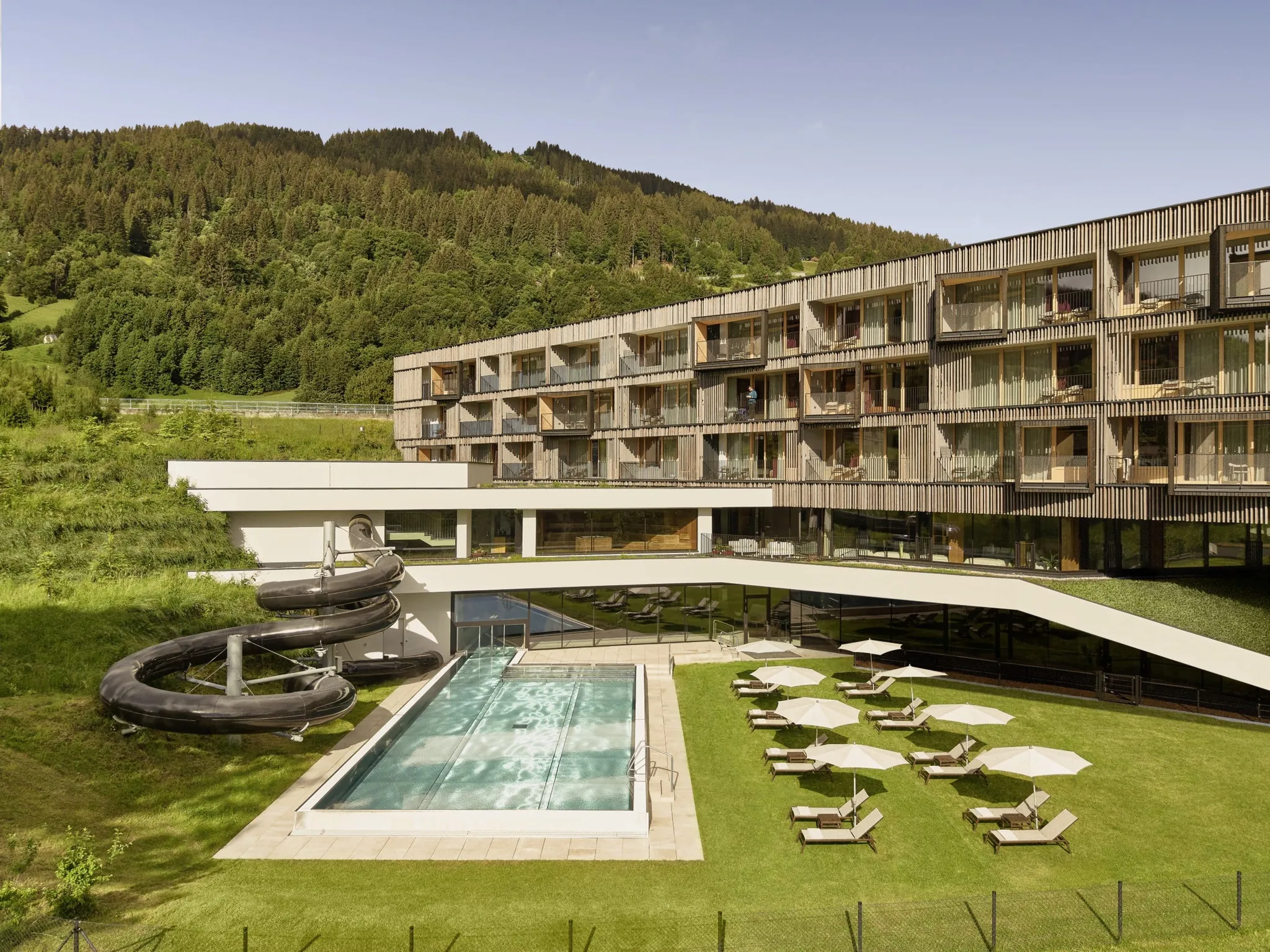 Außenansicht des Falkensteiner Family Hotel Montafon mit Pool, Wasserrutsche und Liegewiese, umgeben von grüner Landschaft