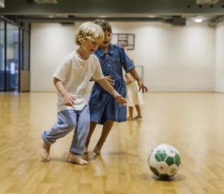 Kinder spielen barfuß Fußball in einer geräumigen Indoor-Halle des Falkensteiner Family Hotel Montafon, während ein Erwachsener im Hintergrund zusieht