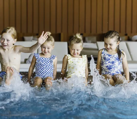 Vier Kinder in Badekleidung sitzen am Rand des Indoorpools im Falkensteiner Family Hotel Montafon und planschen freudig mit den Füßen im Wasser