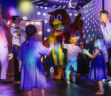 Kinder tanzen mit einem Maskottchen in einer farbenfroh beleuchteten Kinderdisco, begleitet von Erwachsenen im Falkensteiner Family Hotel Montafon