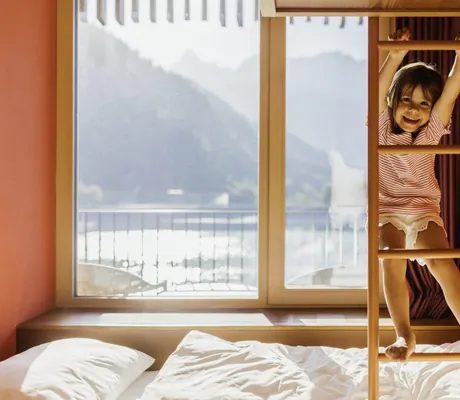 Glückliches Mädchen in einem Zimmer des Falkensteiner Family Hotel Montafon, klettert auf einer Holzleiter vor einem großen Fenster