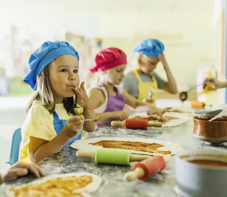 Kinder mit bunten Kochmützen und Schürzen bereiten Pizza zu und probieren die Sauce im Falkensteiner Family Hotel Montafon