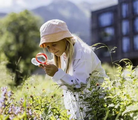 Mädchen mit Sonnenhut und Laborkittel untersucht Pflanzen mit einer Lupe im Freien vor einer Bergkulisse, im Hintergrund das Falkensteiner Family Hotel Montafon
