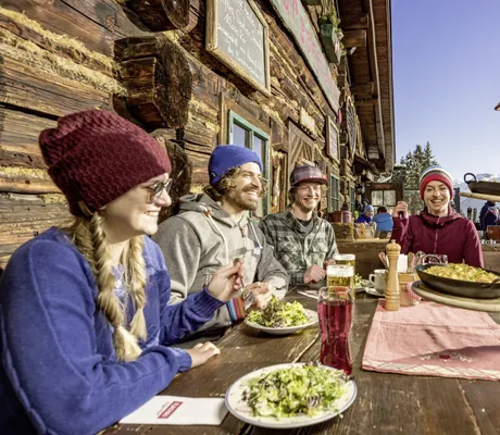 Eine Gruppe Menschen sitzt zusammen vor einer Holzhütte und isst gemeinsam im Freien.