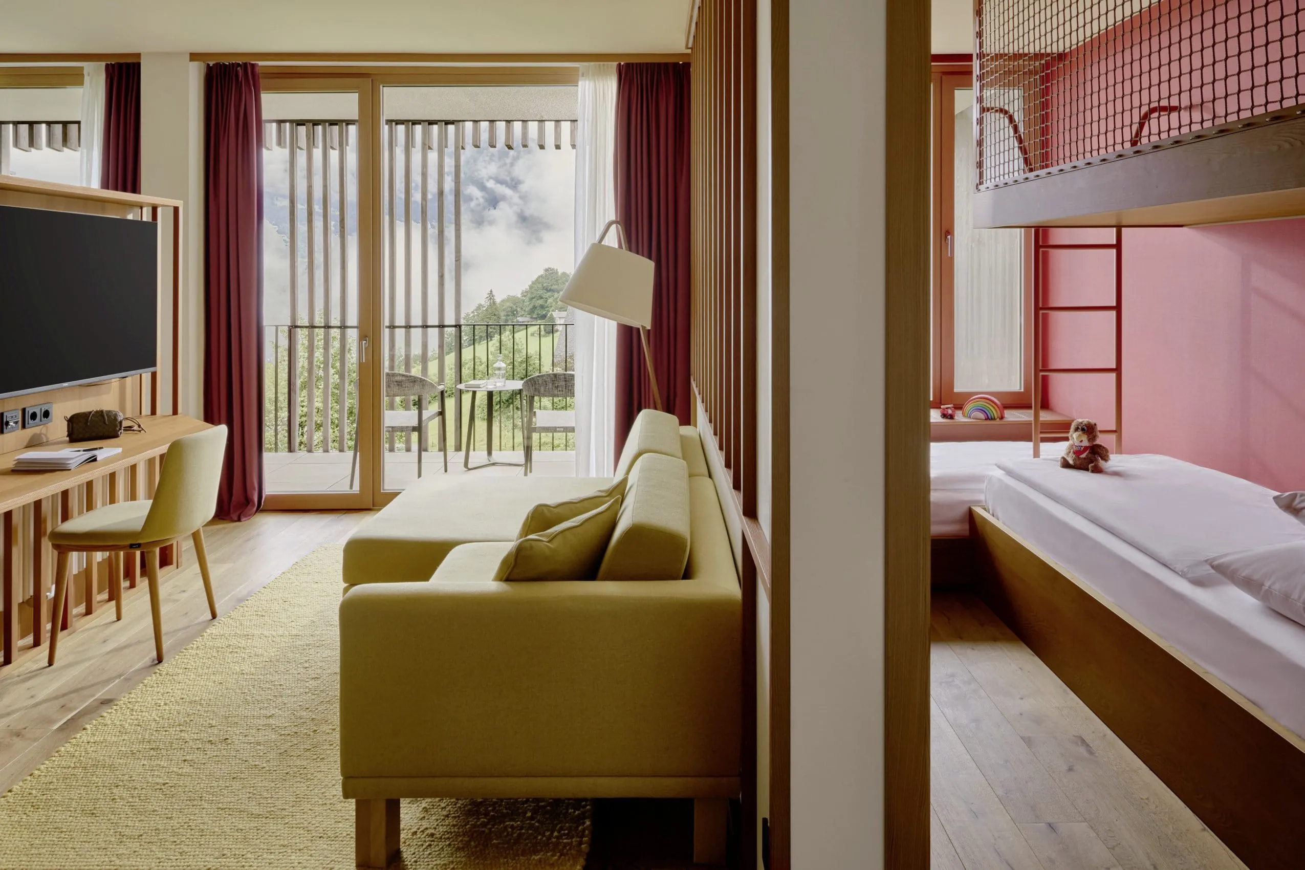 Helle Mountain Suite mit gelb-gr&uuml;nem Sofa, Schreibtisch und Balkonblick auf gr&uuml;ne H&uuml;gel, Kinderbereich mit Hochbett und Spielzeug im Falkensteiner Family Hotel Montafon