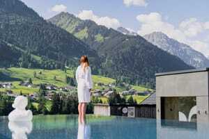 Frau in weißem Bademantel steht am Outdoorpool des Genuss- und Aktivhotel Sonnenburg, bewaldetete Berge im Hintergrund