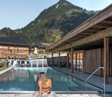 Frau am Rand des Outdoorpools der Alpzitt-Chalets mit Blick auf Chalets und bewaldete Berge im Hintergrund