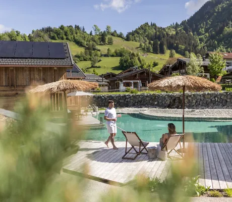 Paar am Naturbadesee der Alpzitt-Chalets mit Holzdeck, Stühlen und Schirmen, eingebettet in eine grüne Berglandschaft