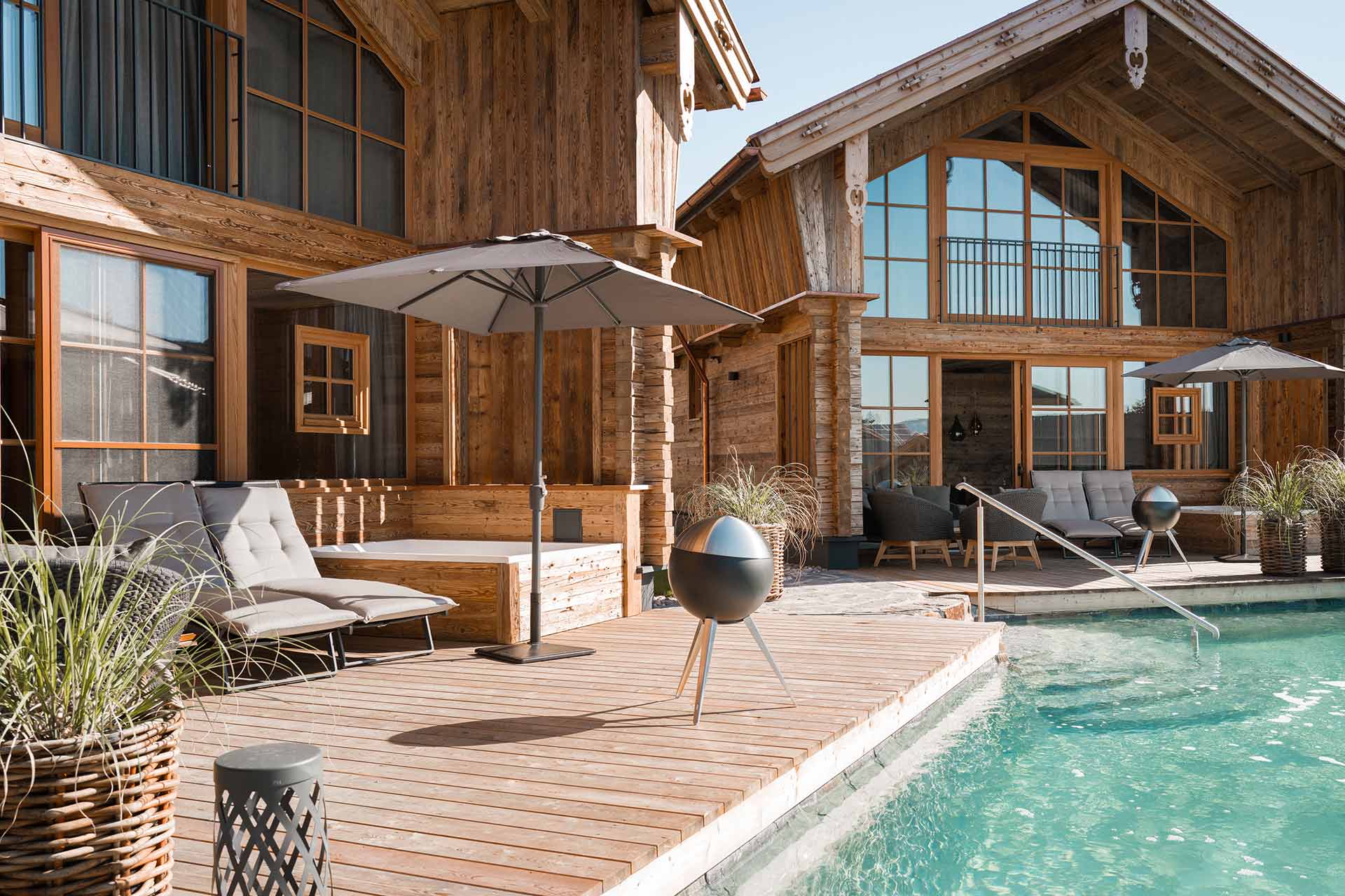 Außenansicht der modernen Alpzitt-Chalets mit Holzterrasse, Liegestühlen und Naturbadeteich in sonniger Umgebung