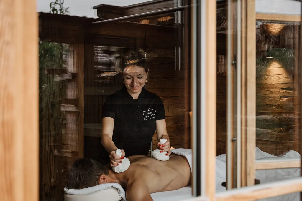 Spa-Behandlung im Spabereich der Alpzitt-Chalets, Therapeutin massiert einen Gast mit Kräuterstempeln, durch ein Fenster sichtbar