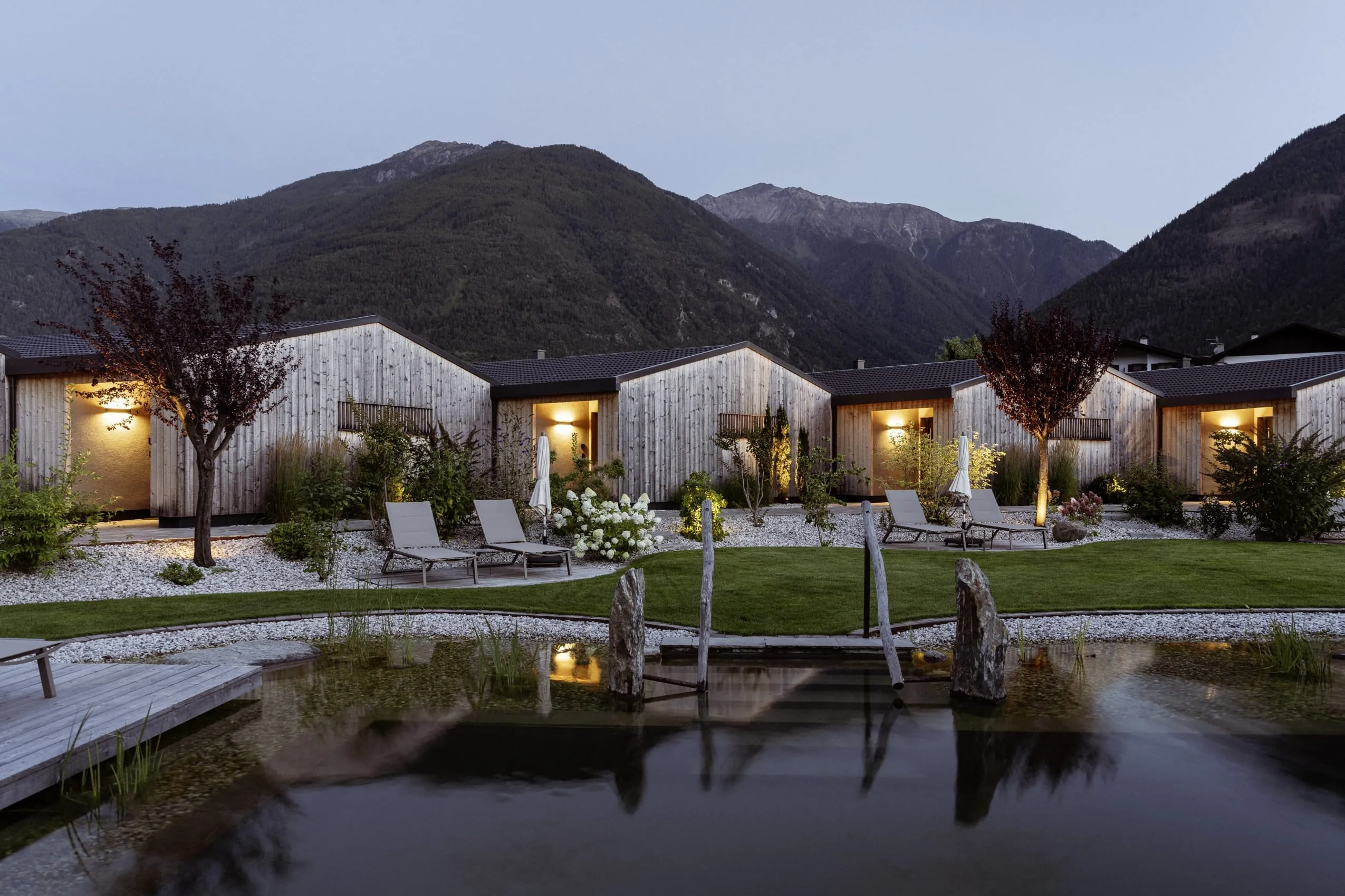 Die Chalets des Amolaris Garden Lodges & Guesthouse bei Dämmerung mit Naturbadesee und Vinschger Bergwelt.