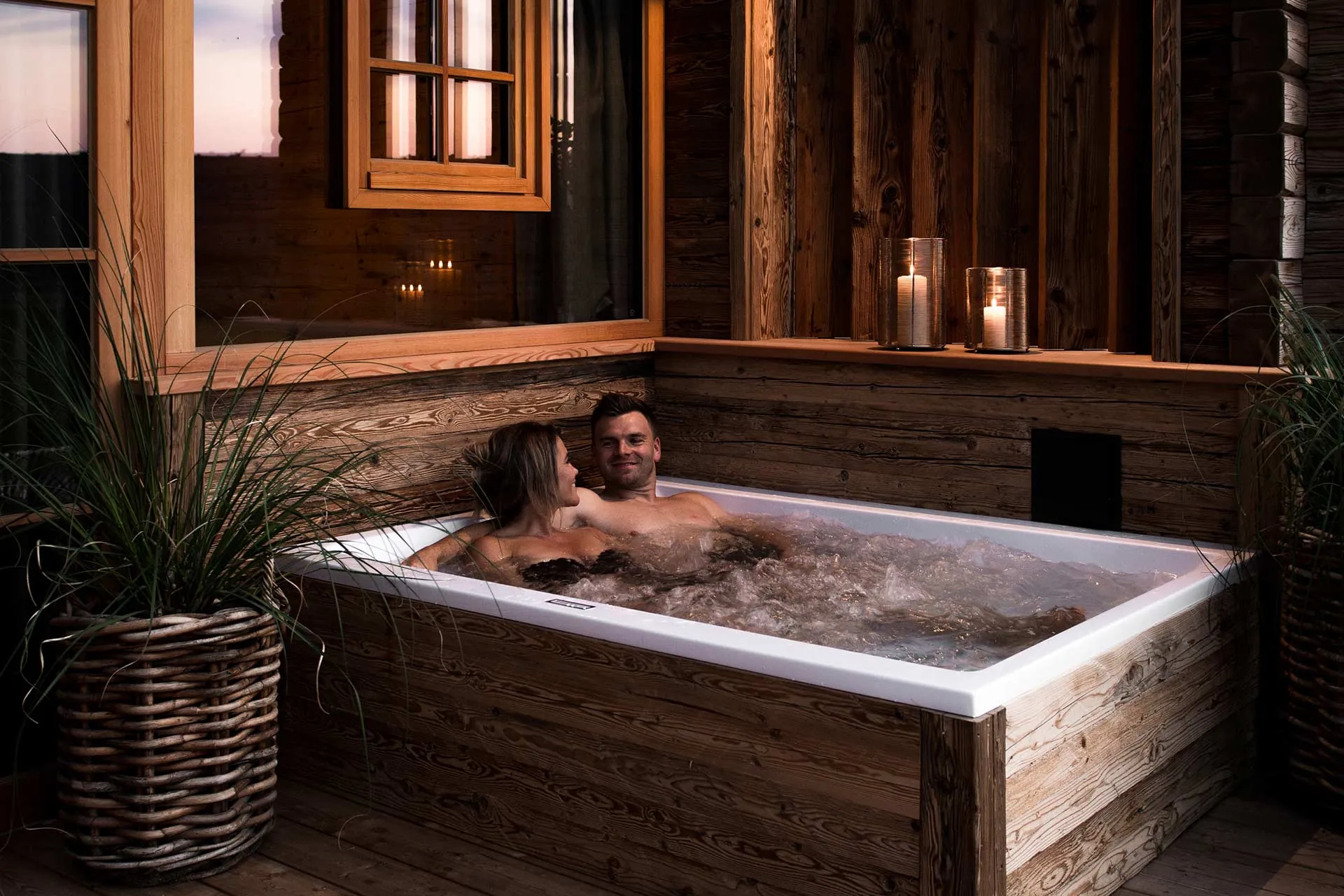 Paar in einer Outdoor-Whirlwanne der Alpzitt-Chalets, umgeben von Kerzen und Pflanzen