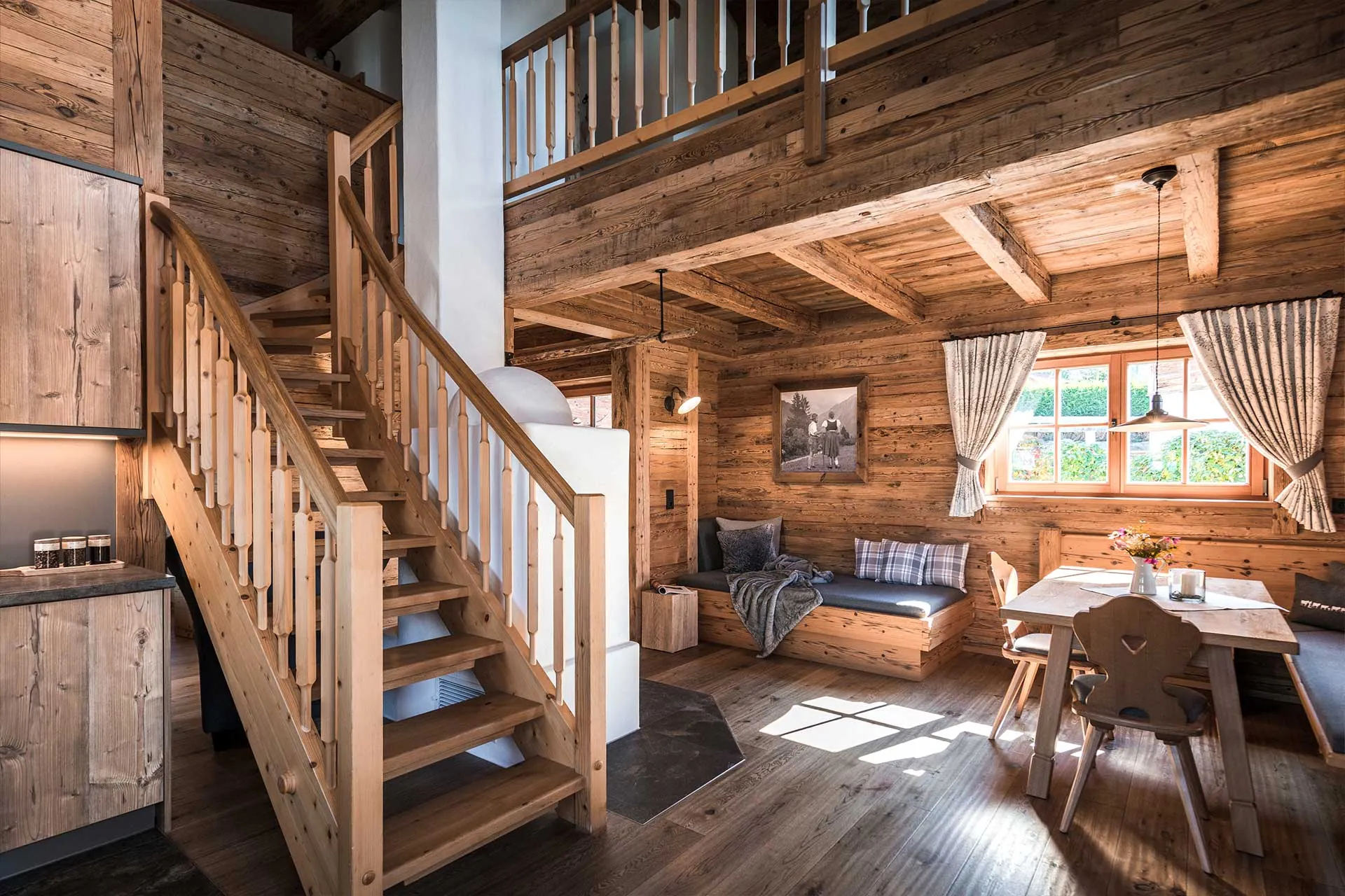 Gemütlicher Wohnbereich eines Alpzitt-Chalets mit Holzvertäfelung, Treppe, Sitzbereich und Esstisch am Fenster