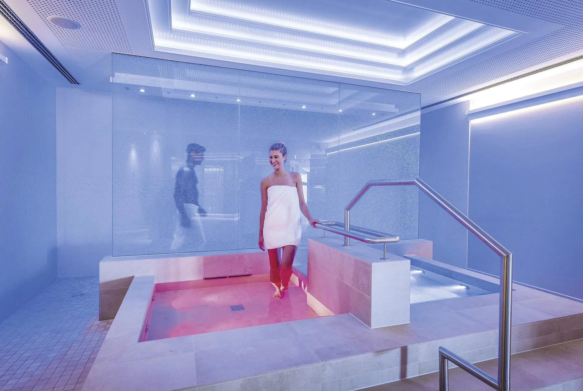 Eine Frau geht durch das Kneippbecken im Innenbereich des Hotel Mooshof Wellness & Spa Resort, w&auml;hrend ein Mann im Wellnessbereich sich erfrischt.