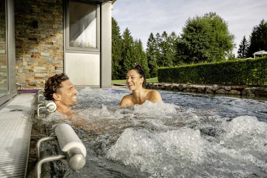 Ein Paar genießt lachend die Wärme des Outdoor-Whirlpools im Hotel Mooshof Wellness & Spa Resort.
