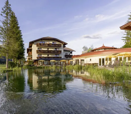 Blick über den Naturteich des Alpenhotel Karwendel umringt von Bäumen auf die Fassade mit Holzbalkonen