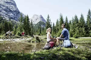 Paar mit Wanderrucksäcken macht auf einem Stein am Bach Pause, während Biker vorbeifahren