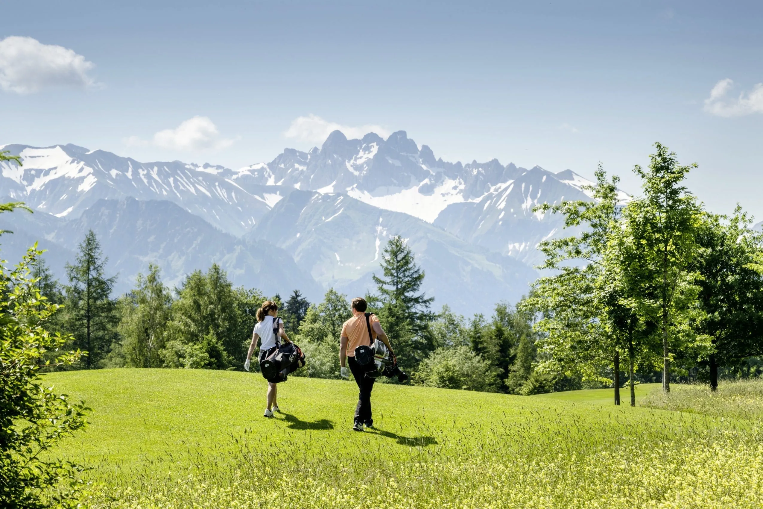 Ein Mann und eine Frau laufen mit Sportausr&uuml;stung &uuml;ber eine gr&uuml;ne Wiese, unweit vom Sonnenalp Resort.
