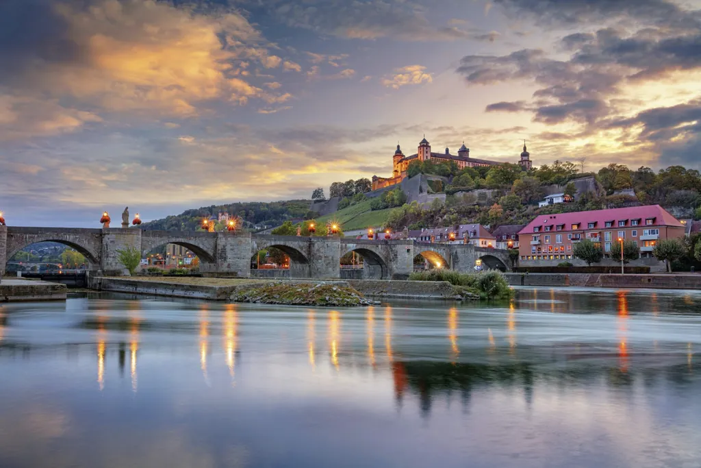 Abendliche Ansicht der Alten Mainbrücke in Würzburg mit beleuchteten Statuen, Wasserreflexionen und der Festung Marienberg im Hintergrund